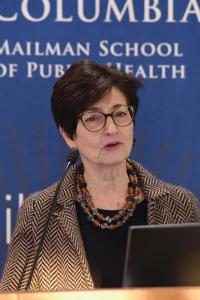 A woman wearing glasses speaks at a podium in front of a Columbia backdrop
